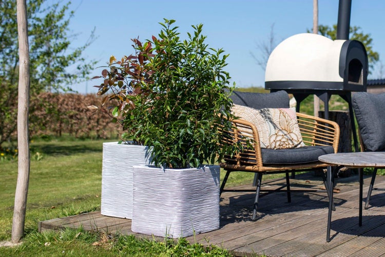 Helle Gartenszene mit zwei quadratischen, strukturierten Pflanzkübeln, einem Korbsessel mit Kissen und einem Pizzaofen auf einer Holzterrasse.