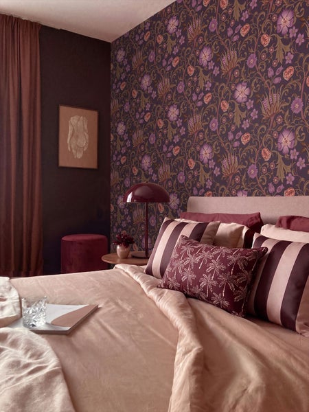 Schlafzimmer mit floraler Mustertapete, einer dunklen Wand, einem Bett mit gemusterten Kissen und einem Beistelltisch mit Lampe.