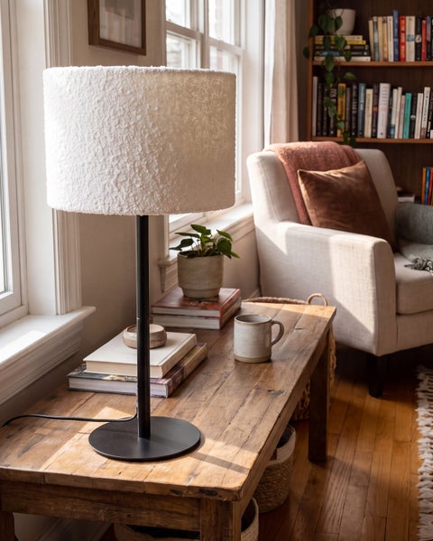 Moderne Tischlampe mit strukturiertem Lampenschirm auf einem Holztisch in einer gemütlichen Leseecke mit Sessel und Bücherregal.