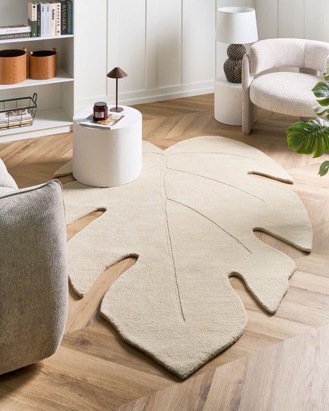 Ein helles Wohnzimmer mit einem beigen Teppich in Form eines Monstera-Blattes auf einem Holzfußboden in Fischgrätoptik.