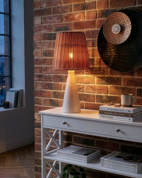 Tischleuchte mit Rattanschirm und Keramikfuß auf weißem Konsolentisch vor einer Backsteinwand.