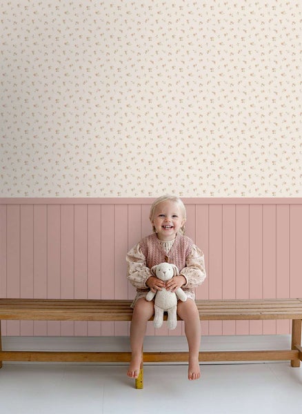 Kindersitzbank aus Holz mit einem lachenden Kind und Stoffhase vor einer Wand mit Tapete mit Blumenmuster und unterem Paneel in Holzoptik.