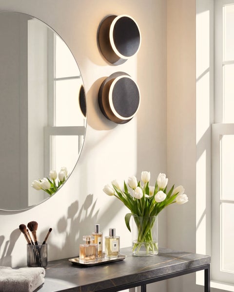 Heller Schminkbereich mit zwei runden LED-Wandleuchten, rundem Spiegel und einem Marmortisch mit weißen Tulpen und Parfümflaschen.