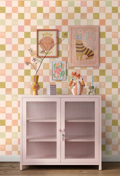 Einrichtungsbeispiel mit karierter Tapete in Pastelltönen, einem rosa Metallschrank mit Glastüren und farbenfroher Wanddekoration.