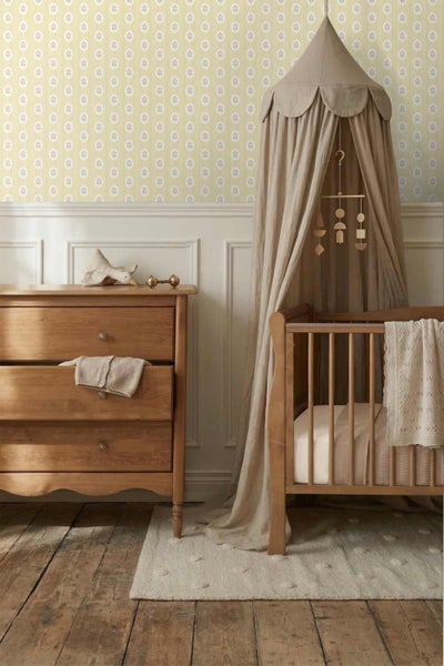 Helles Kinderzimmer mit einer Holzkommode, einem Gitterbett aus Holz mit Stoffhimmel und gemusterter Tapete an der Wand.