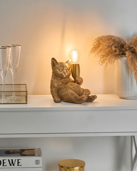 Goldfarbene Tischleuchte in Form einer sitzenden Katze, die eine Glühbirne hält, auf einer weißen Anrichte dekoriert.