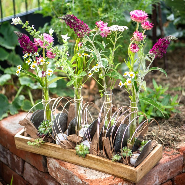 Dekoratives Holztablett mit vier Glasvasen in einem Drahtgestell, arrangiert mit Gartenblumen, Rindenstücken, Schieferplatten und Sukkulenten.