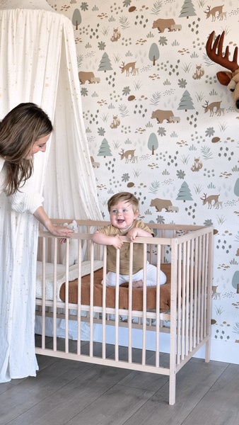 Ein helles und freundliches Kinderzimmer mit einem Baby im Gitterbett, dekoriert mit einer Waldtapete und einem Baldachin.