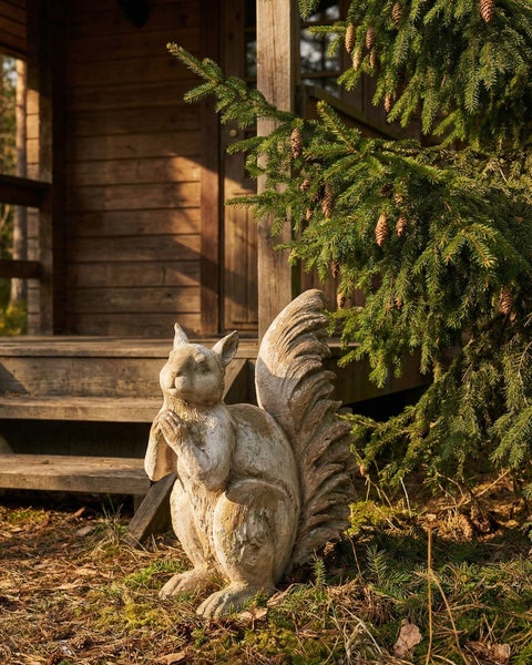 Gartenfigur Eichhörnchen aus Steinoptik, sitzend vor einer Holzhütte im Garten neben einem Nadelbaum.