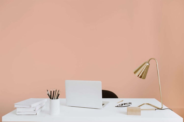 Moderner Arbeitsplatz mit weißem Schreibtisch, Laptop und goldener Schreibtischlampe vor einer gestrichenen Wand. Hell und organisiert.