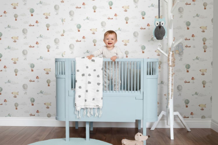 Baby im blauen Kinderbett vor einer Tapete mit Flugmotiven