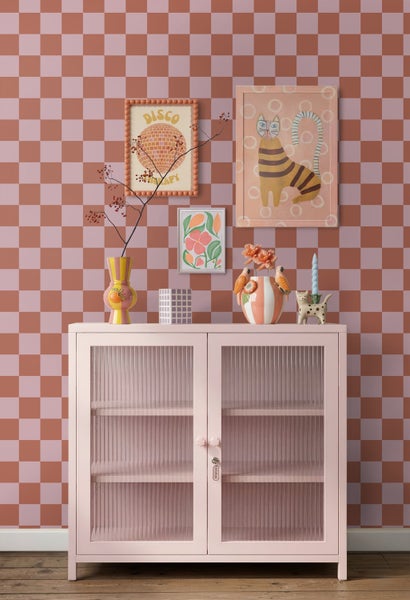 Wandgestaltung mit karierter Tapete in Terrakotta und Rosa, davor eine rosa Kommode mit Glastüren und verschiedenen dekorativen Vasen und Bildern.