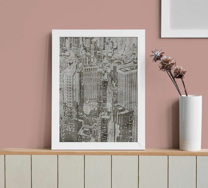 Gerahmtes Wandbild mit schwarz-weißer Illustration einer Stadtsilhouette auf einem Holzregal neben einer weißen Vase mit Trockenblumen.