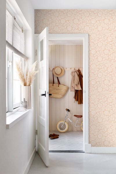 Hell gestalteter Raum mit floral gemusterter Vliestapete, Blick durch eine weiße Tür in einen Flur mit Holzpaneelen und Laufrad.
