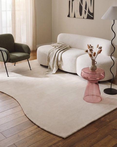 Moderner Wohnraum mit hellem, geschwungenem Teppich, weißem Sofa, grünem Sessel, rosa Beistelltisch und schwarzer Stehlampe auf Holzboden.