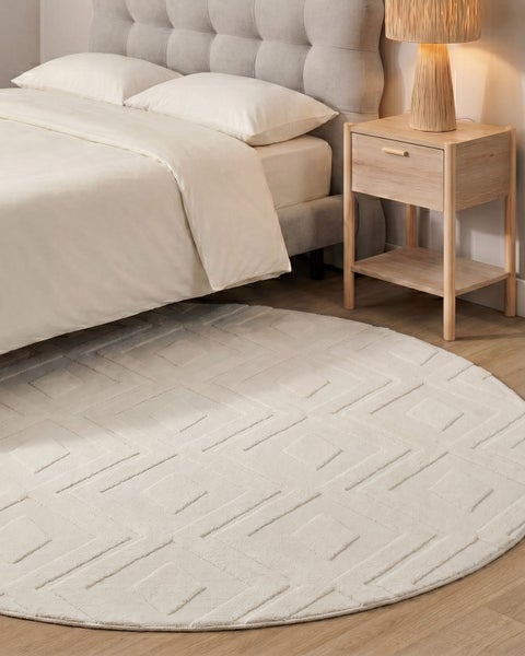 Runder Teppich mit geometrischem Muster vor einem gepolsterten Bett mit Nachttisch und Tischlampe in einem modern eingerichteten Schlafzimmer.