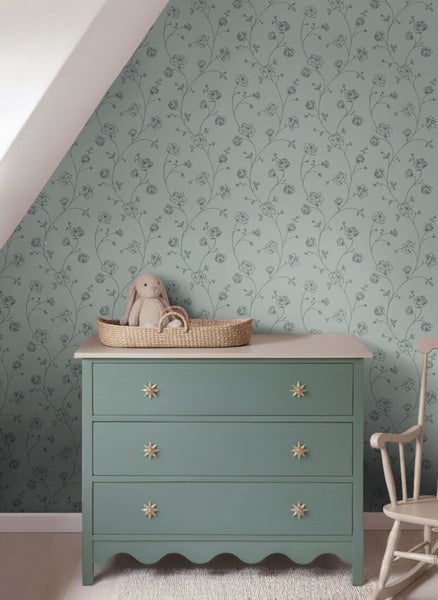 Kinderzimmer mit floral gemusterter Tapete, Kommode mit Sterngriffen, Weidenkorb mit Stofftier und hölzernem Schaukelstuhl.