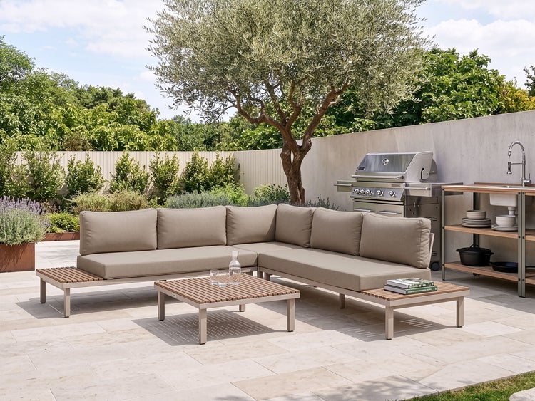 Moderne Loungegruppe mit Ecksofa und Couchtisch auf einer Steinterrasse neben einer Außenküche mit Grill und Spülbecken im Garten.