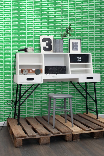 Schreibtisch mit Aufbewahrungselementen, schwarzen Metallbeinen und einem grauen Hocker auf einer Holzpalette vor einer grünen Tapete mit Automotiven.