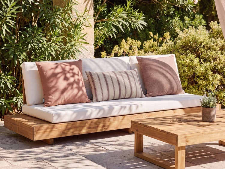 Gartensofa aus Holz mit hellen Sitzpolstern und dekorativen Zierkissen auf einer Terrasse im Garten bei sonnigem Wetter.