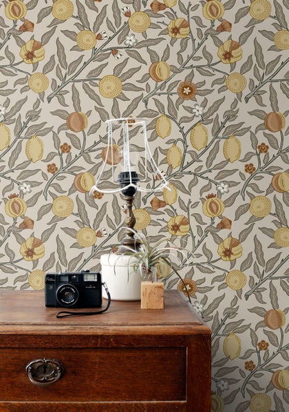 Eine Nahaufnahme einer Kommode mit einer Kamera, einer Lampe und einer Pflanze vor einer Tapete mit einem Muster aus Zitronen, Pfirsichen, Blumen und Blättern.