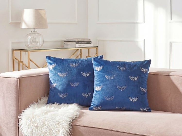 Zwei dunkelblaue Samtkissen mit Schmetterlingsmuster auf einem Sofa in einem hellen Wohnzimmer.