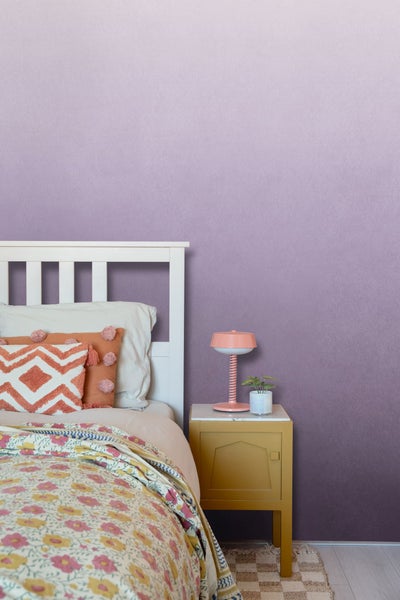 Schlafzimmer mit Bett, Nachttisch und Lampe vor einer lila Wand