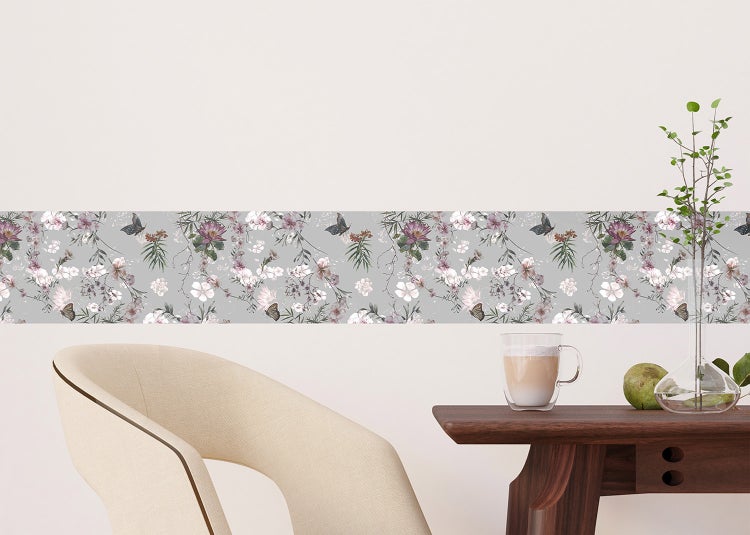 Selbstklebende Bordüre mit Blumen- und Schmetterlingsmuster an einer Wand über einem Holztisch mit Kaffeetasse und Stuhl.