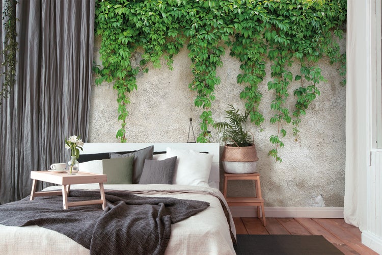 Fototapete mit grünen Kletterpflanzen auf einer hellen Wand in Steinoptik hinter einem Bett im Schlafzimmer.