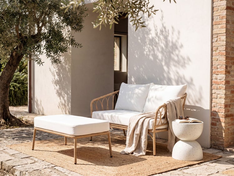 Garten-Lounge-Set aus Rattan mit weißen Polstern und Hocker auf einer mediterranen Terrasse mit Olivenbaum und Naturteppich.