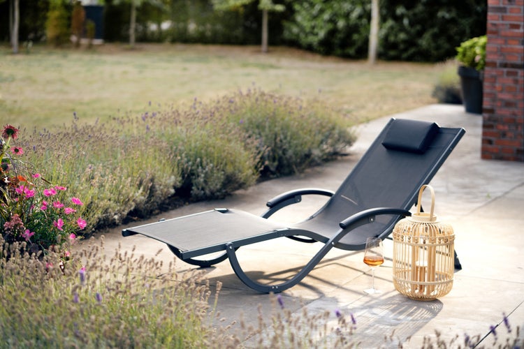 Gartenliege mit Kopfstütze auf einer Terrasse neben einem Windlicht und Lavendelblüten im Garten.