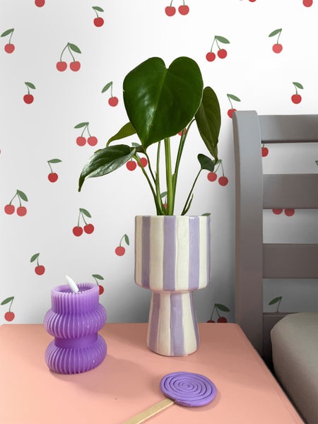 Szene mit Zimmerpflanze in gestreiftem Blumentopf, gewellter Kerze und Kerzenlöscher auf einem Tisch.