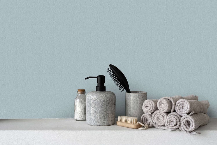 Hellblaue strukturierte Tapete in einem Badezimmer mit Seifenspender, Haarbürste und aufgerollten Handtüchern auf einer weißen Ablage.