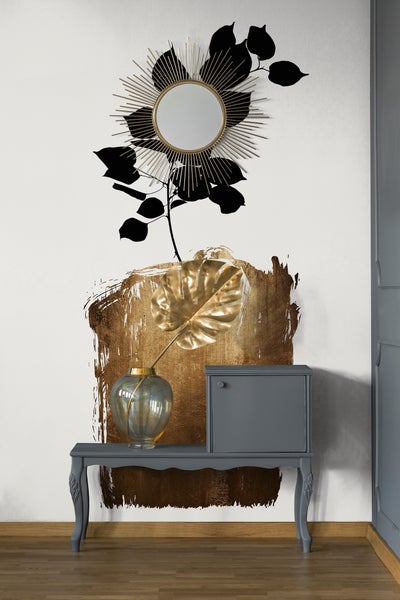 Wandgestaltung mit goldenem Pinselstrich, schwarzem Pflanzenmotiv und Sonnenspiegel über einer grauen Konsole mit Monstera-Blatt in einer Vase.