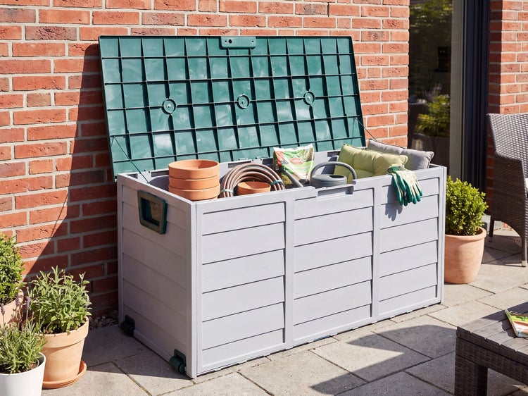 Geöffnete Gartenbox aus Kunststoff mit verstautem Gartenbedarf auf einer Terrasse vor einer Ziegelwand.