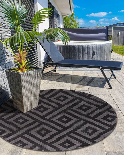 Moderner Außenbereich mit rundem Outdoor-Teppich mit geometrischem Muster, Sonnenliege, Pflanzgefäß mit Palme und rundem Whirlpool auf Steinterrasse.