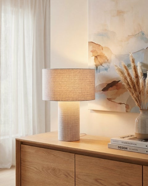 Moderne Tischleuchte mit Schirm und Fuß in Leinenoptik auf einem Sideboard aus Holz in einem hellen Wohnraum neben Büchern und einer Vase.
