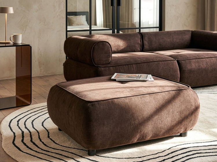 Modernes Wohnzimmer mit braunem Polsterhocker und passendem Sofa auf einem gemusterten Teppich vor einem Beistelltisch mit Lampe.