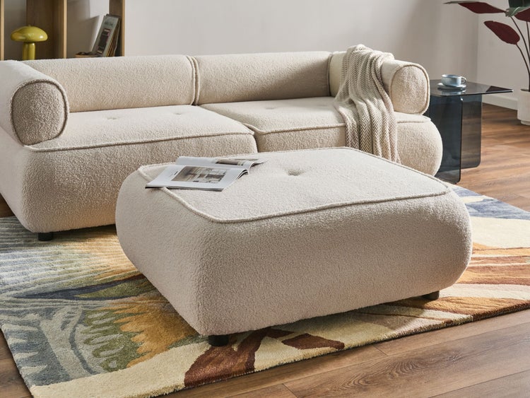 Quadratischer Hocker aus Bouclé-Stoff in einem modern eingerichteten Wohnzimmer mit passendem Sofa auf einem gemusterten Teppich.