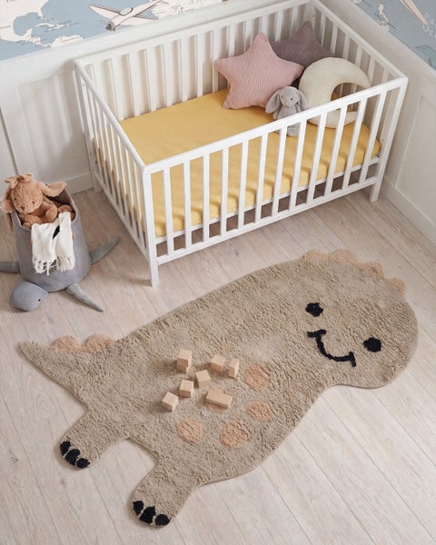 Ein Kinderzimmer mit einem weißen Babybett, einem Dinosaurier Spielteppich und Holzbausteinen auf dem Boden.