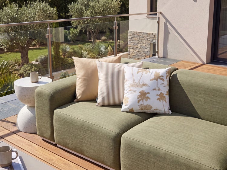 Modernes Lounge Sofa auf einer Holzterrasse mit drei Zierkissen, kombiniert mit einem runden Beistelltisch vor einer Glasbrüstung im Freien.