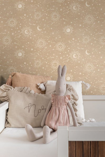 Kindertapete mit Sonnen, Monden, Sternen und Wolken in einem hellen Kinderzimmer mit Kissen und Stoffhase auf dem Bett.