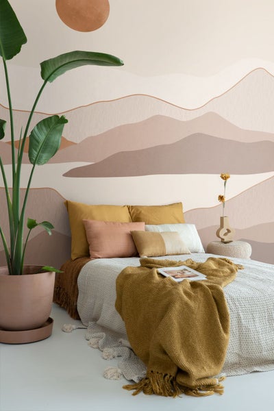 Schlafzimmer mit Wandbild Berglandschaft in Erdtönen, Bett mit Kissen und Strickdecke sowie großer Zimmerpflanze im Tontopf.