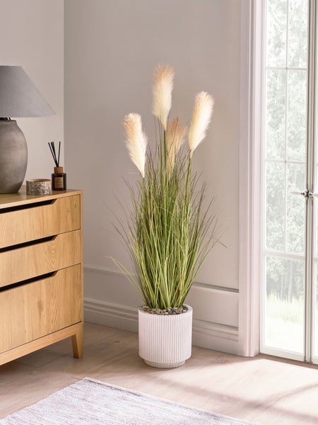 Dekorative Kunstpflanze mit Pampasgras in weißem Topf neben einer Kommode