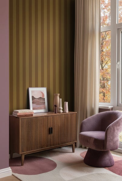 Modern eingerichtetes Zimmer mit gestreifter Tapete, einem Holzsideboard mit Rillenfront, einem Sessel und langen Vorhängen am Fenster.