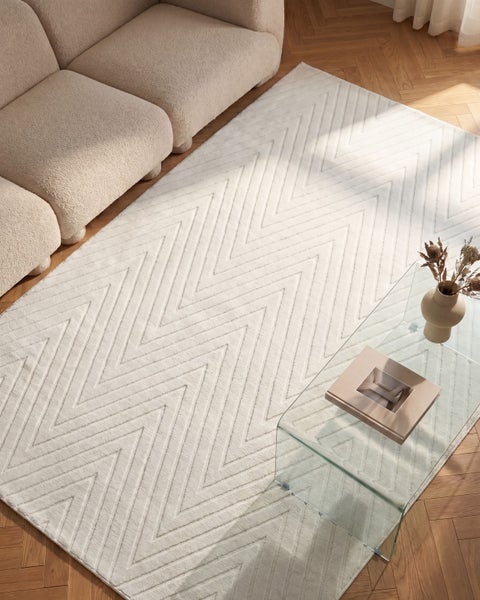 Ein rechteckiger Teppich mit Zickzack Muster liegt vor einem Sofa auf einem Parkettboden in einem modern eingerichteten Wohnzimmer.