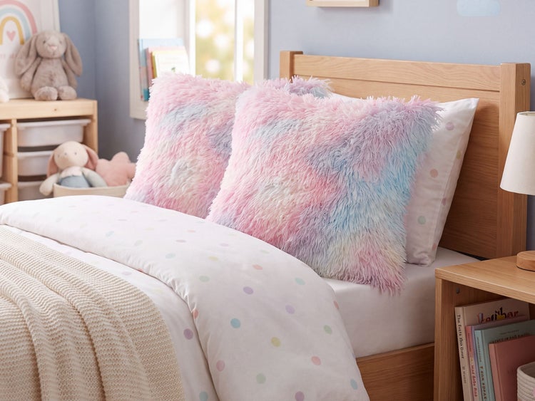 Zwei flauschige Zierkissen mit Langflor in Pastellfarben auf einem Kinderbett mit gepunkteter Bettwäsche und Holzgestell.