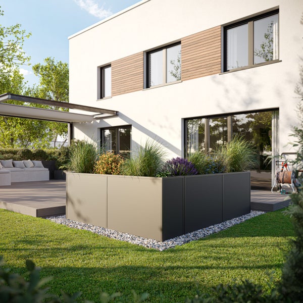 Modernes L-förmiges Hochbeet aus Metall mit Gräsern und Blumen auf einer Rasenfläche neben einer Terrasse und einem hellen Haus.