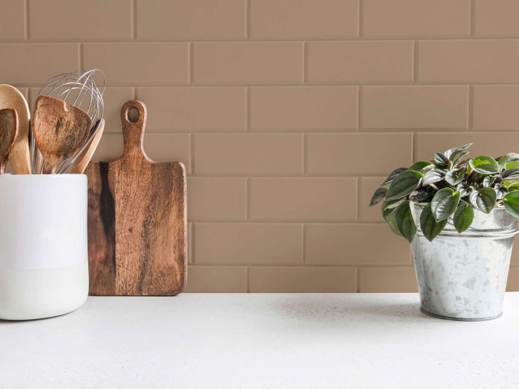 Küchenrückwand mit rechteckigen Wandfliesen in Beige-Braun, davor eine weiße Arbeitsplatte mit Küchenutensilien und einer Topfpflanze.