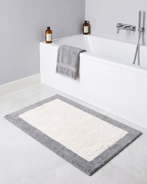 Rechteckiger Bademattenvorleger mit texturierter Mitte und kontrastierendem Rand vor einer modernen Badewanne mit Duscharmatur im Badezimmer.